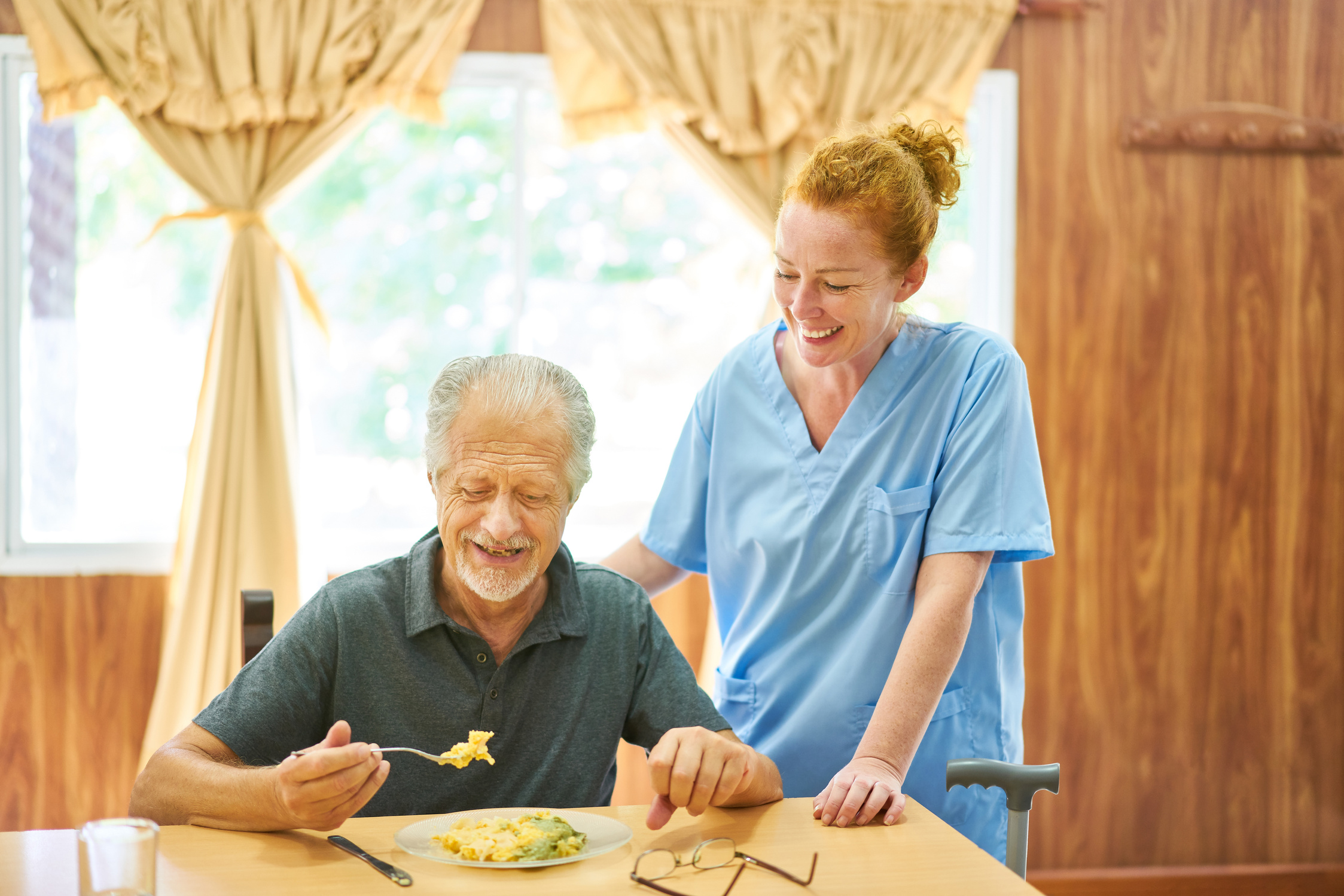 An older man eating scrambled eggs at a breakfast table. A younger nurse is smiling and assisting him.