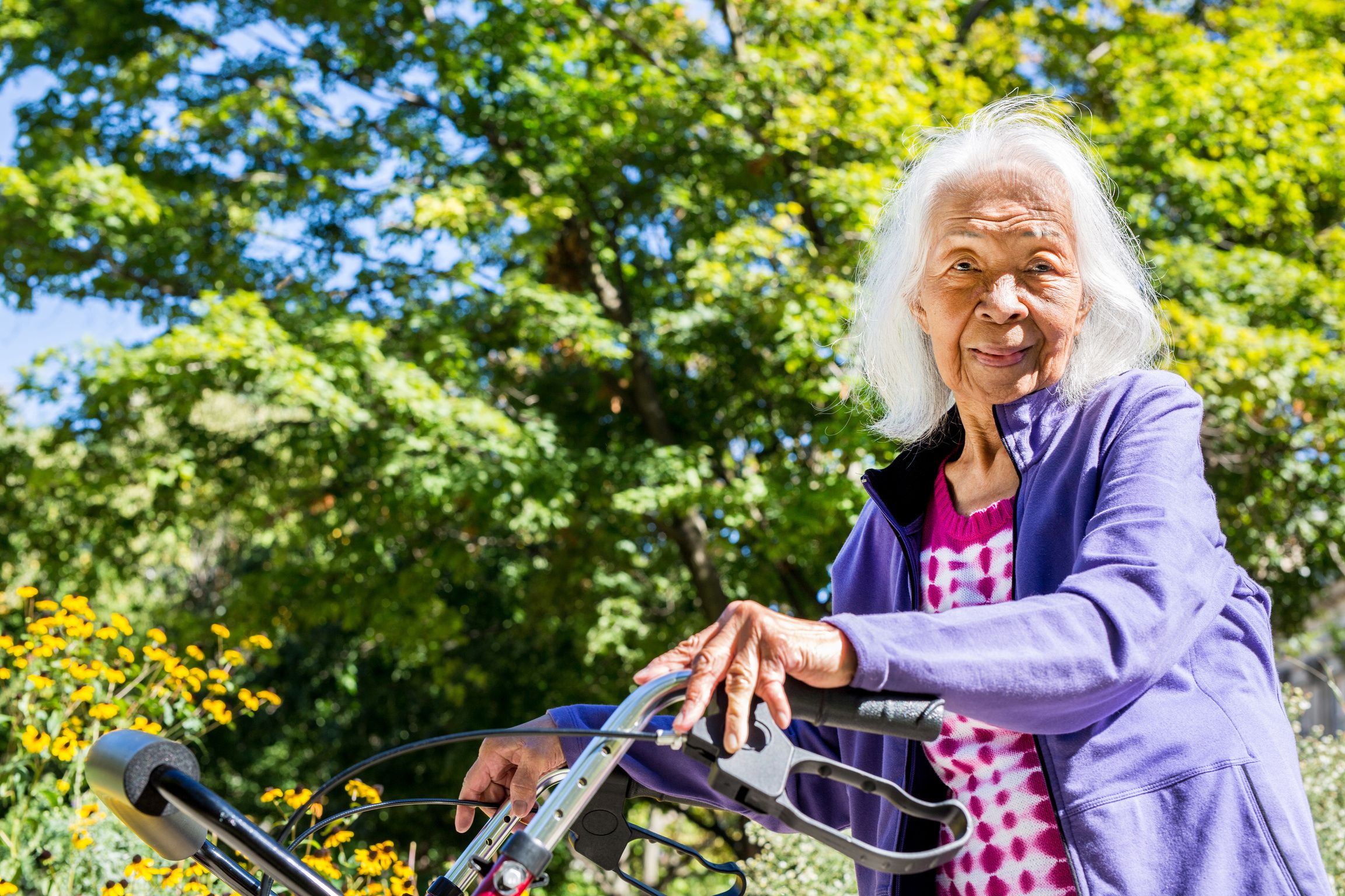 An old woman wearing a lavender color coat using a walking assistance device smiling at the camera. There are trees and flowers behind her.