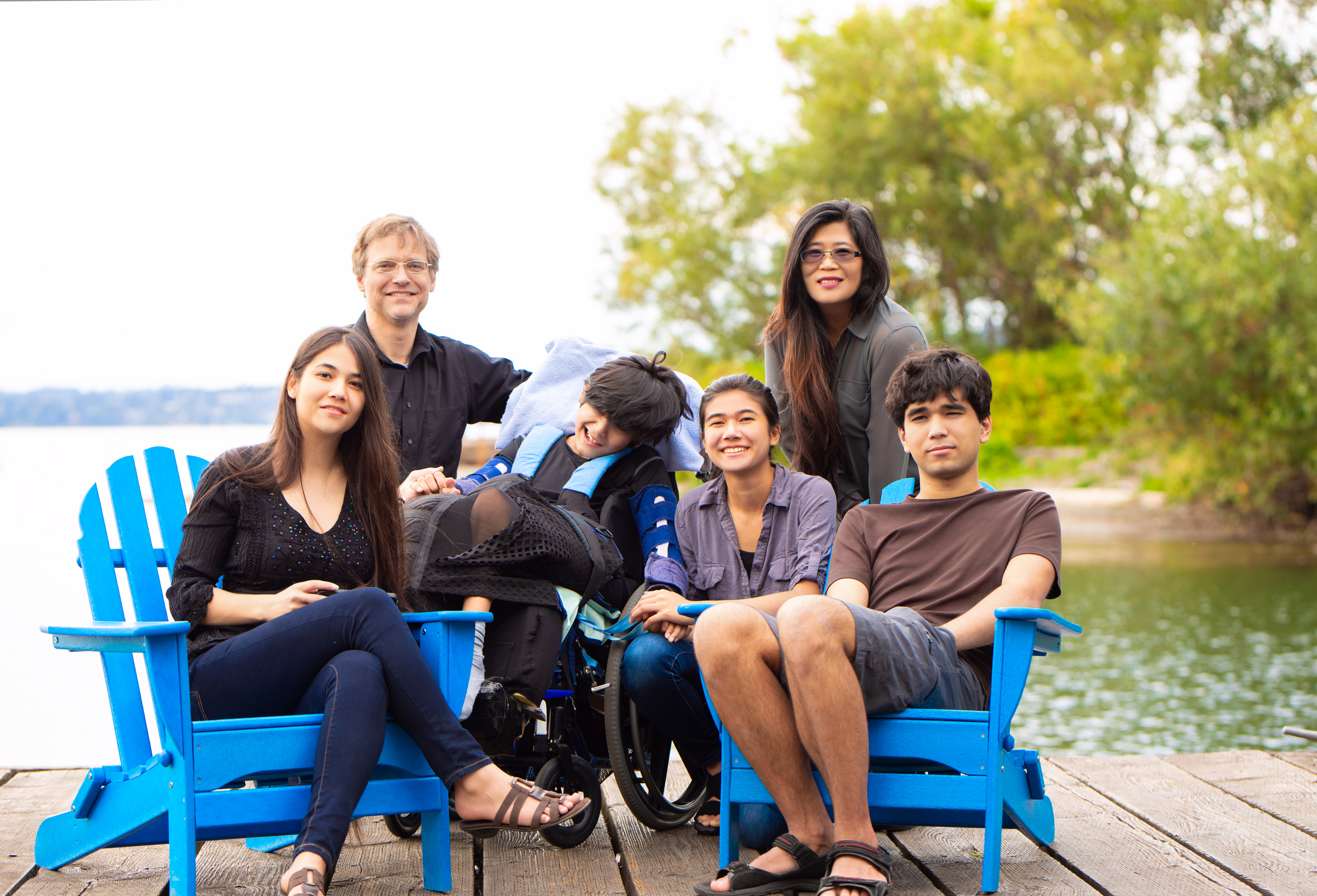 A family sitting on blue chairs on a dock by a lake. All members are smiling. The young man in the middle is using a wheelchair.