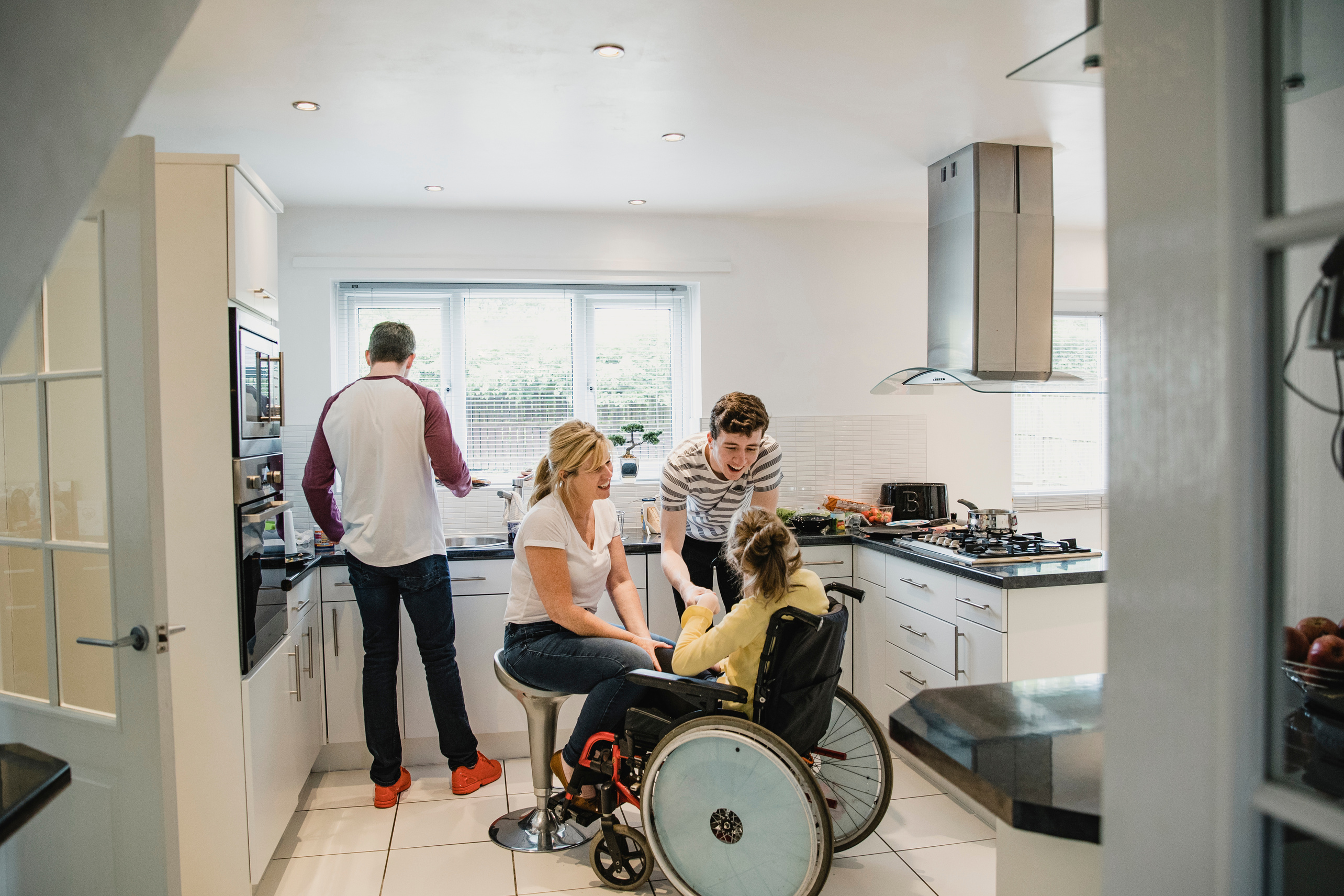 A young woman using a wheelchair shaking hands with a smiling young man. An older woman sits on chair in front of her, also smiling. A man is washing dishes.