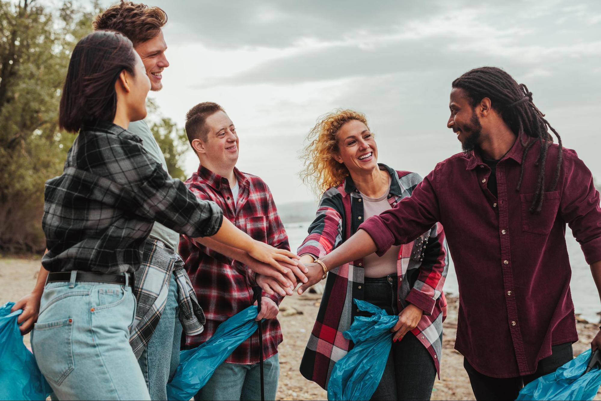 Five people on a beach are standing in a circle with their hands in the middle. They are carrying bags to help pick up trash. One of the men has a developmental disability.