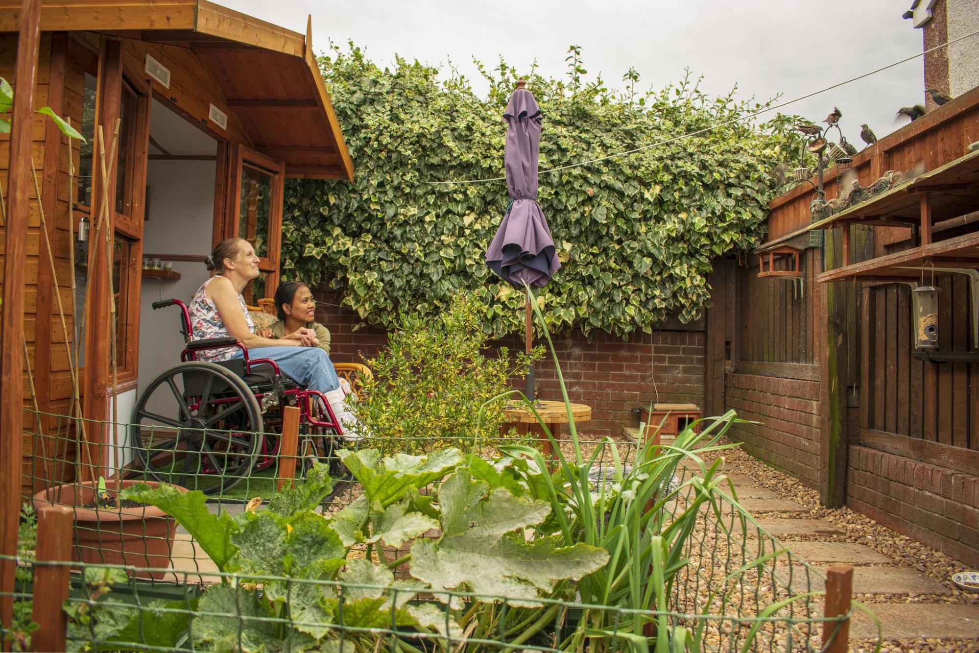 Two women are admiring a small urban backyard with lots of leaves in the background and lettuce growing in the foreground. One of the women is in a wheelchair.