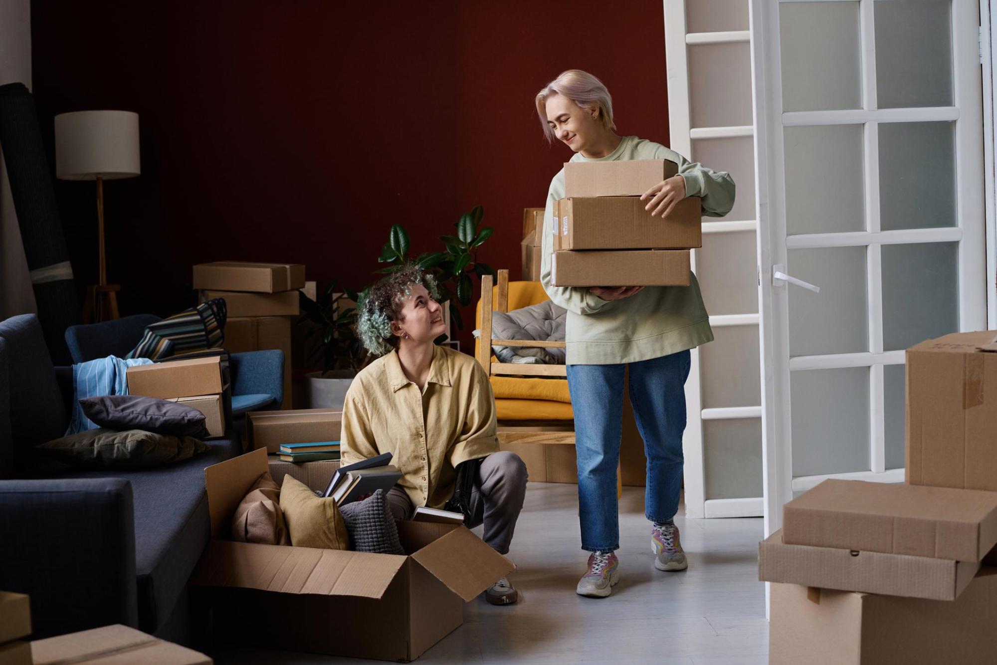 Two women are moving boxes into a cozy new living space with white floors and dark red walls . One of the women has a prosthetic arm.