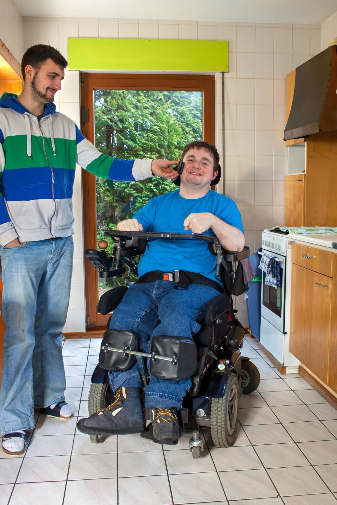 Two men are in a kitchen. One man smiling is in a wheelchair wth accessibility equipment for his hands. The other man is helping the first man hold a phone to his ear.