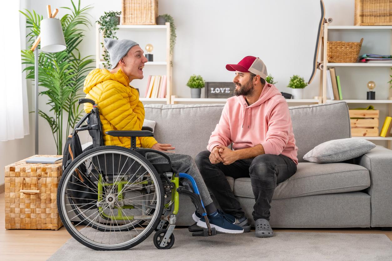 A man in a wheel chair and a winter coat is conversing with another man who is sitting on a couch. They appear to be laughing.