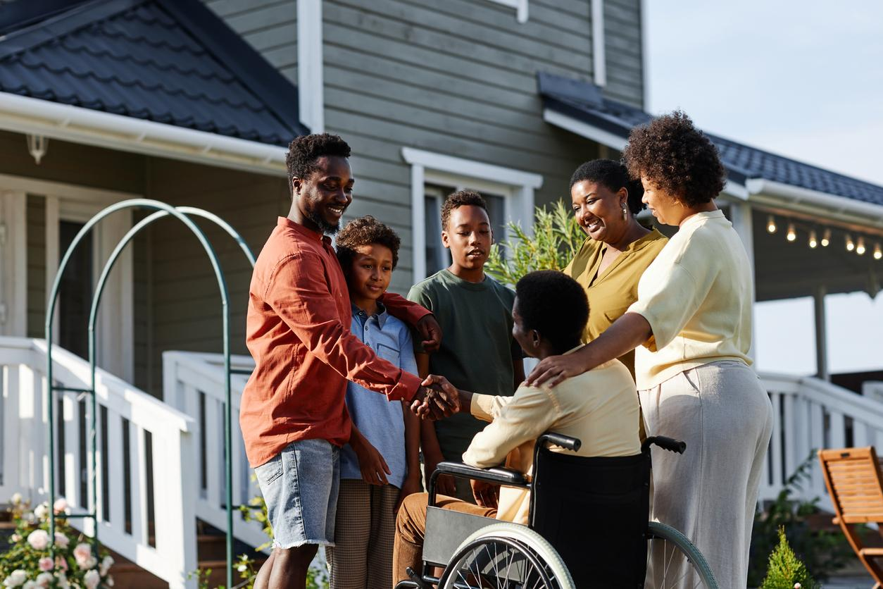 A man in a wheelchair is surrounded a by five members of his family. He's shaking hands with the a man in the group while a woman has her arm around his shoulder. They are in front of a residence and they appear happy.