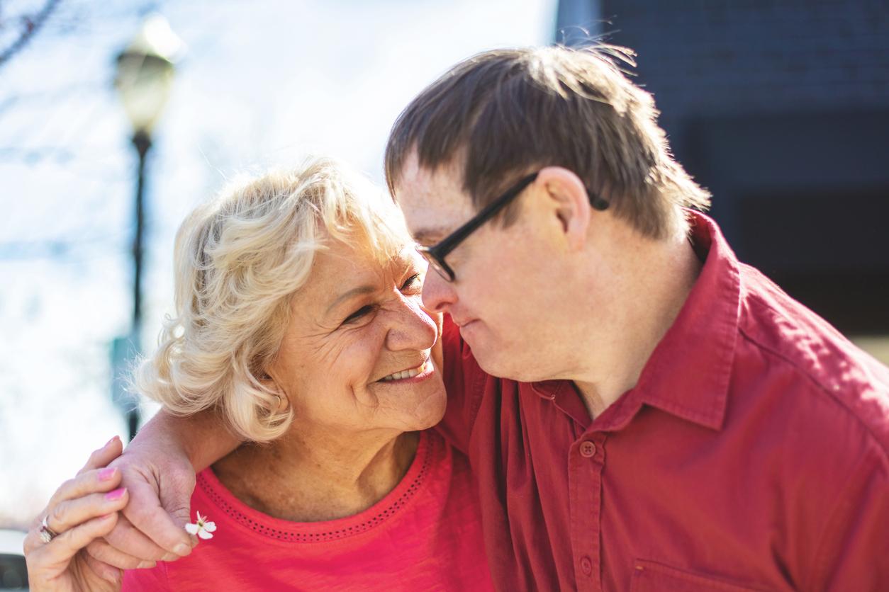 A young man with a disability hugs and older woman who is smiling at him. He is holding a small flower.