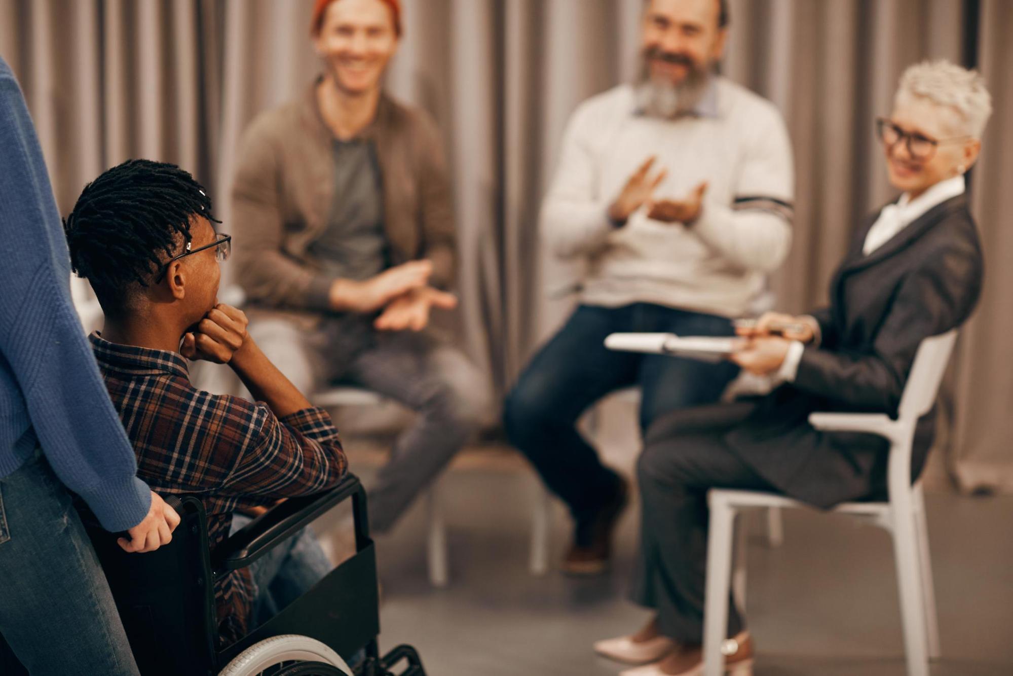 A man in a wheelchair is attending a meeting. There are three people seated around him out of camera focus, but they appear to be applauding for the man in the wheelchair.